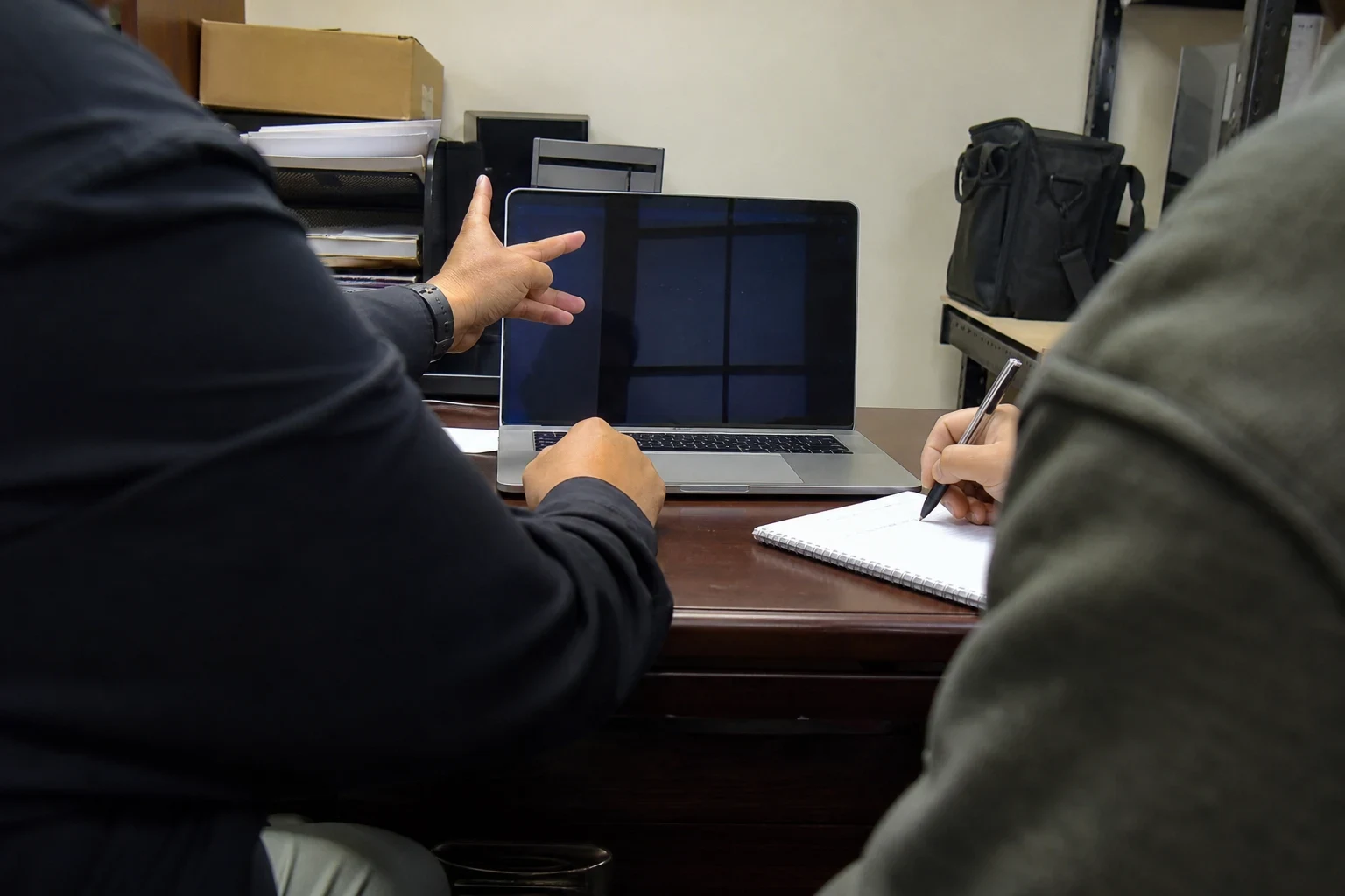 Over-the-shoulder view of two people at a wooden office desk, one pointing at a laptop while the other takes notes in a spiral notebook