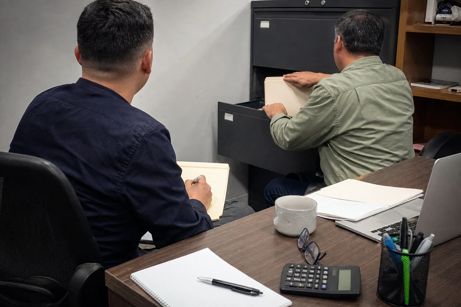 Two men in an office: one seated with a legal pad takes notes while the other pulls a file from a black filing cabinet; a desk holds a calculator, notebook, mug and laptop.