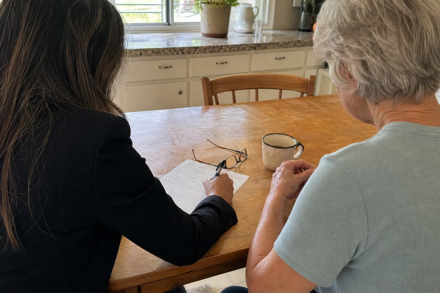 Agent in a dark blazer writing on a one-page form at a wooden kitchen table beside an older woman, with reading glasses and a coffee mug on the table and a kitchen window behind