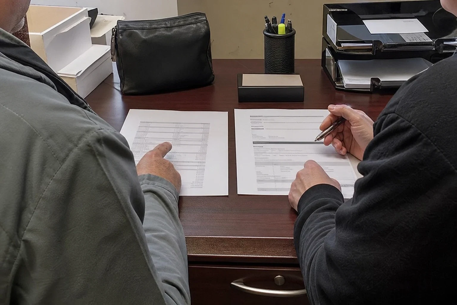 Two people seen from behind at a wooden office desk, reviewing two printed documents side by side, one pointing at figures while the other holds a pen