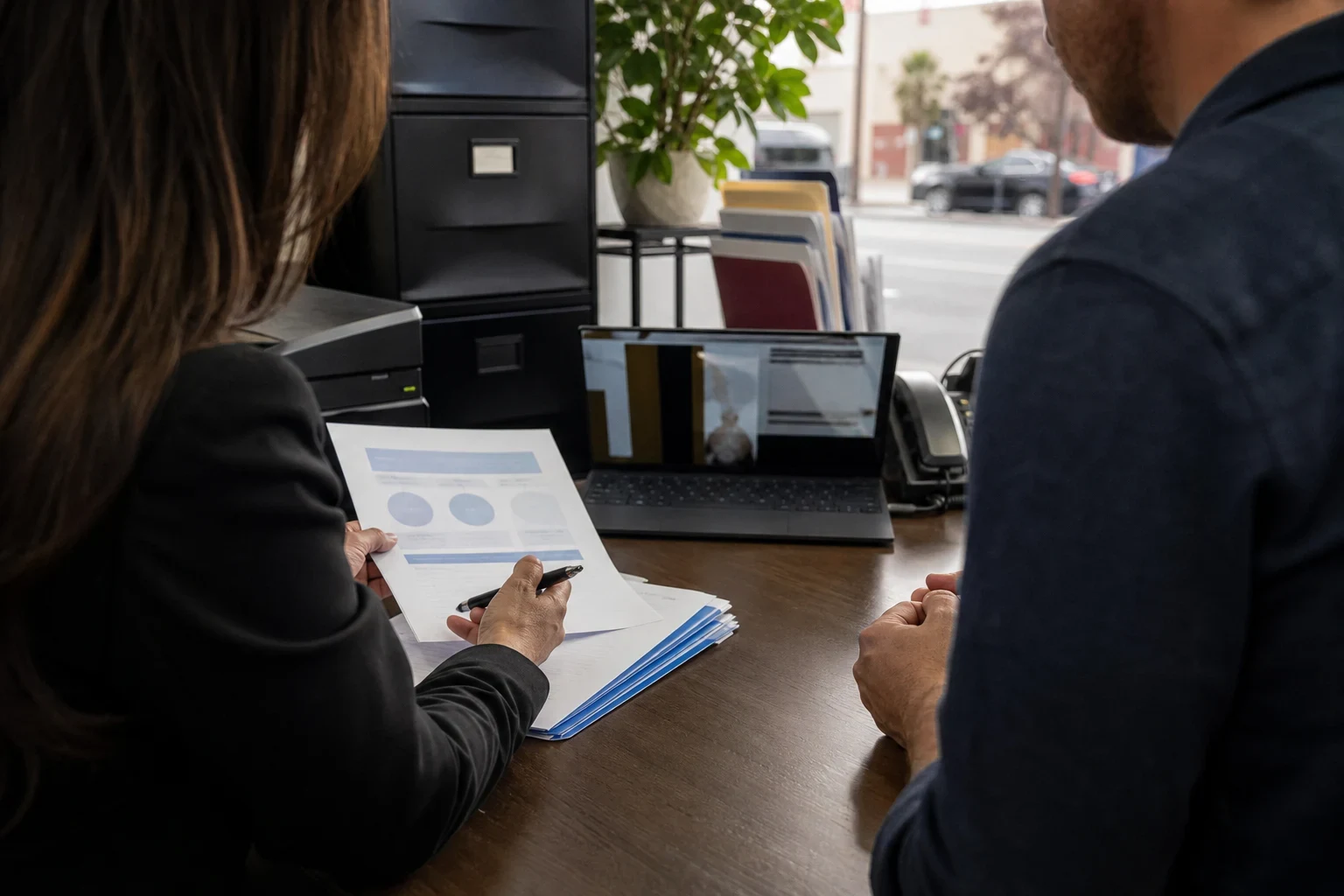 Two people at a wooden office desk reviewing a printed summary beside a laptop, filing cabinet and plant, with a window onto a street and parked cars